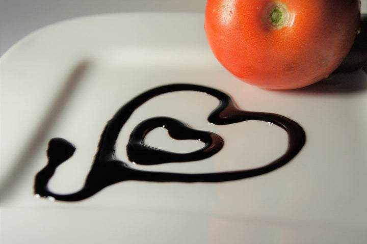 Balsamic vinegar drizzled into shape of a heart on a white plate with a red tomato in the background
